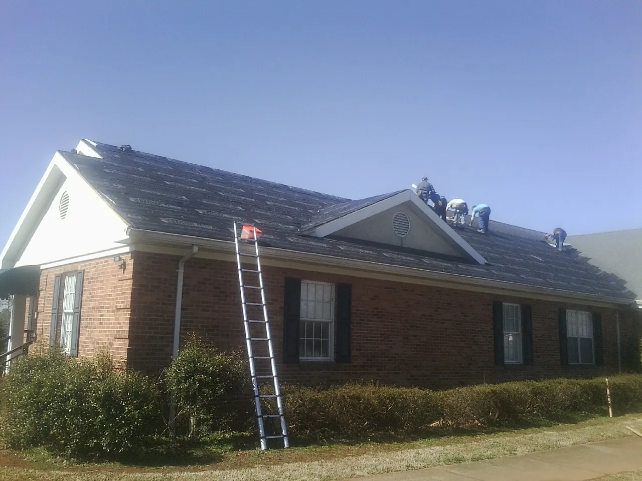 Crew tearing shingles off a brick ranch roof with a ladder set — Metal Roof Repair work by our crew in Westville